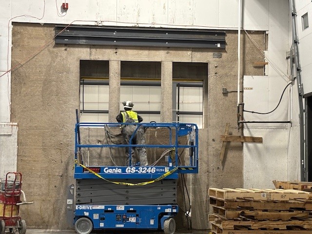 A construction worker in a safety vest and hard hat stands on a blue Genie scissor lift inside an industrial warehouse, working on a large concrete wall with three vertical openings. Stacks of wooden pallets are piled nearby, and construction materials are visible in the background.