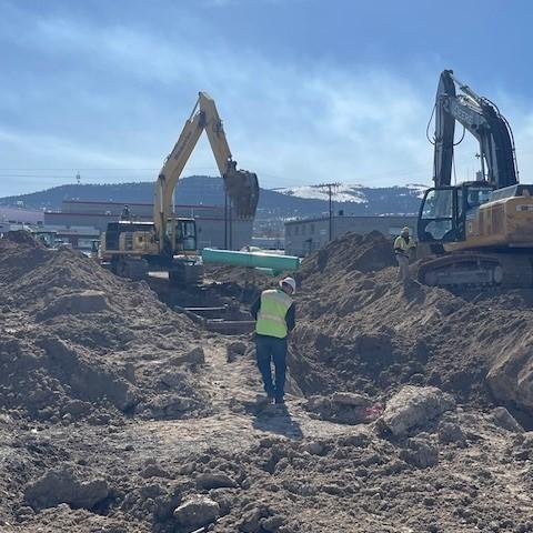 A construction worker wearing a high-visibility vest stands between two large excavators on the liquor warehouse worksite.