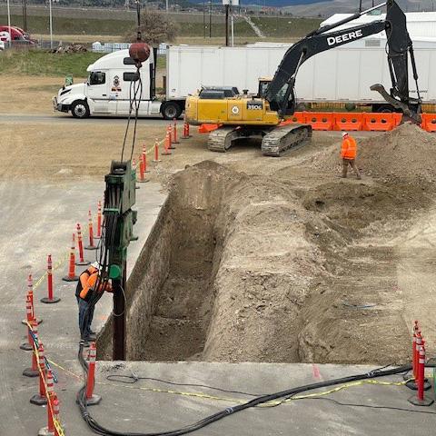 Close-up of a construction worker wearing an orange vest and helmet, working next to a large excavation pit with a heavy drill machine inserting a piling into the ground. Construction barriers and equipment are visible around the site.