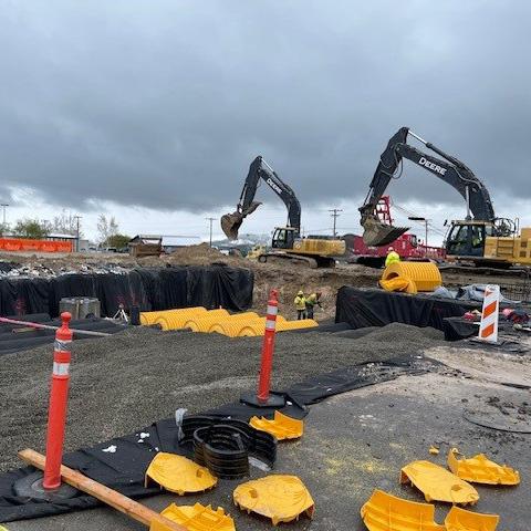 Overcast sky above a construction site with two large excavators digging in a fenced-off area filled with dirt, construction materials, and yellow corrugated pipes laid on the ground.