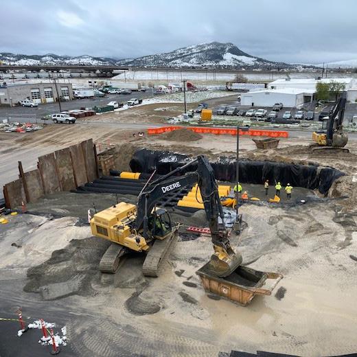 Elevated view of the liquor warehouse construction site featuring heavy machinery, including a bulldozer and an excavator working around a large, deep excavation with protective barriers and construction workers visible.