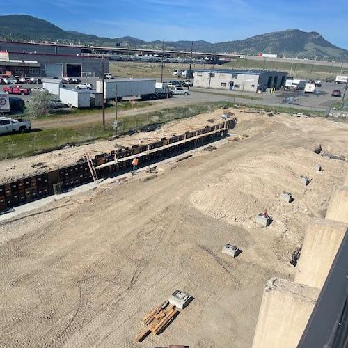 A long trench reinforced with wooden and steel beam footings with scattered construction materials and machinery, lined with the beginnings of the foundation walls.